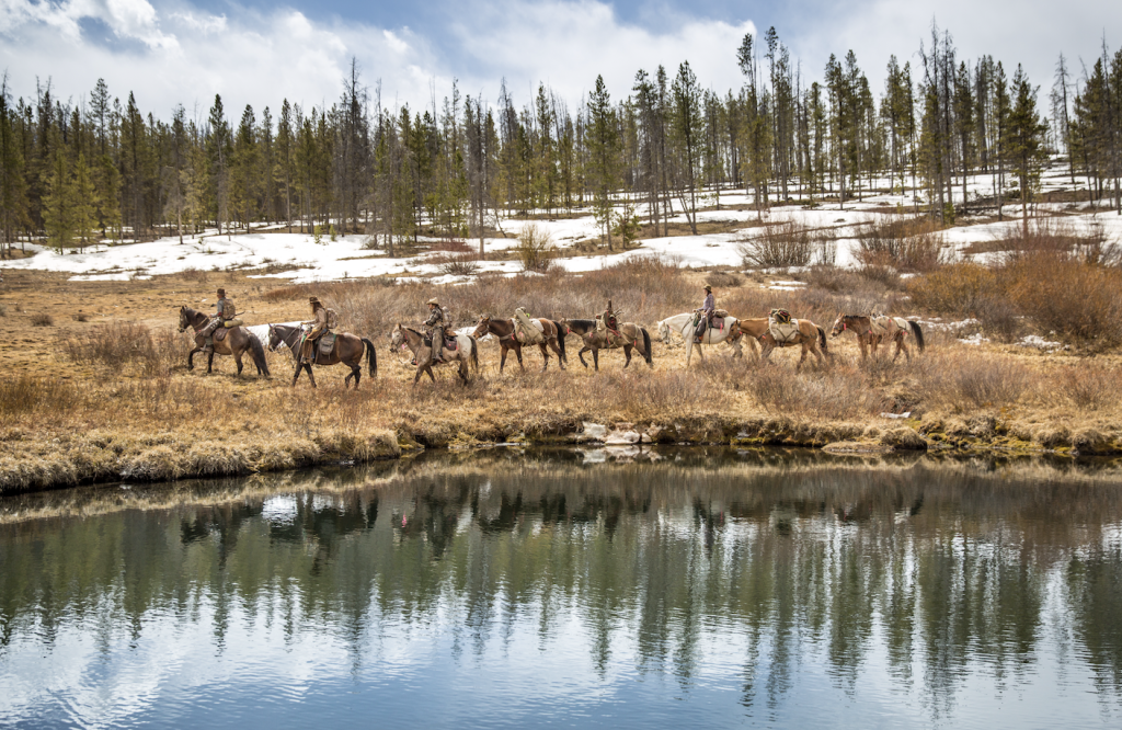 A group of horseback riders traverse a snowy, forested landscape near a calm lake in Grand County Colorado.